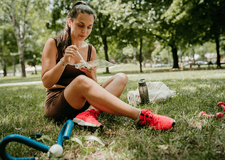 A woman eating a meal from a container while sitting in the grass outside. She's wearing workout clothes.