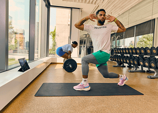 Exerciser deadlifting barbell in background, with another exerciser doing a twist lunge while taking a Peloton App class on a tablet in foreground