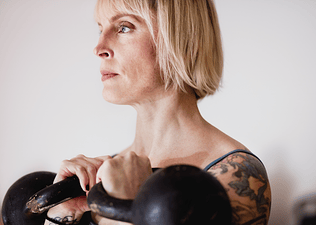 Woman with tattoos holding two kettlebells racked at shoulder height while doing kettlebell shoulder exercises