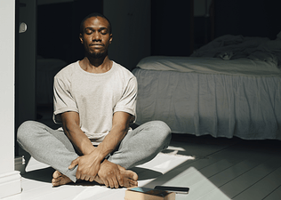 A man calmly sitting on the floor and meditating in his bedroom. Check out eight meditation tips in this article.