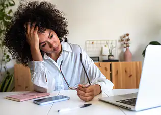 A mentally exhausted woman sitting at her desk in front of her laptop. She's holding her head in one hand and her glasses in another. She looks stressed out and tired.
