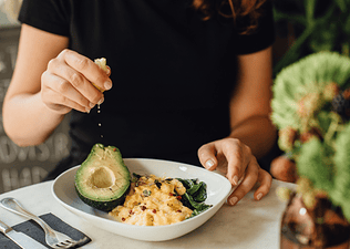 A woman eating scrambled eggs and half an avocado for a meal. She's squeezing a lemon slice on the avocado.