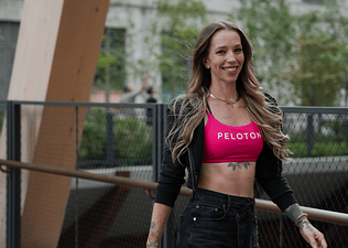 Peloton instructor Kirra Michel going on a walking meditation outdoors. She's smiling and wearing a pink Peloton sports bra, a black zip-up hoodie, and black jeans.