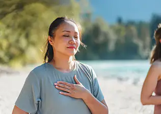 A woman practicing self-compassion meditation outside, softly smiling and holding her hand to her heart.