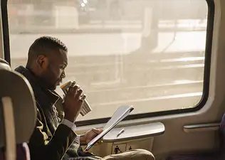 A businessman sipping coffee from a thermos while reading documents while sitting on a sunny train.