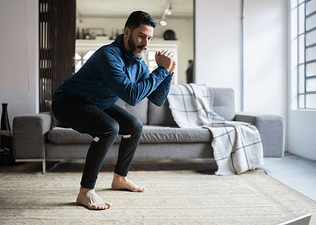 A middle-aged man performing a squat exercise while following a guided workout at home.