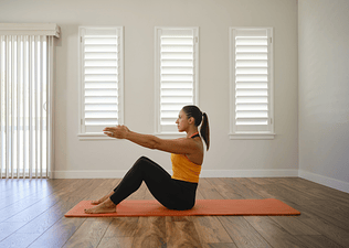 Woman doing a modified Pilates roll-up with bent knees in front of windows on an exercise mat