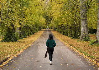 A woman walking down a road surrounded by trees in autumn. Learn the mental health benefits of walking in this article.