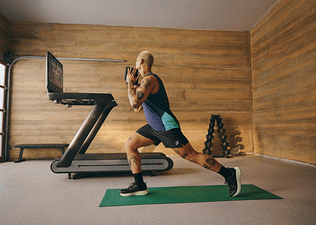 Man doing dumbbell front lunges as part of a quad workout using the Peloton Tread+. 