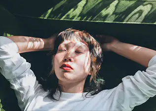 A woman relaxing at home and practicing progressive muscle relaxation. She's lying on a sunny sofa with her eyes closed and hands behind her head.