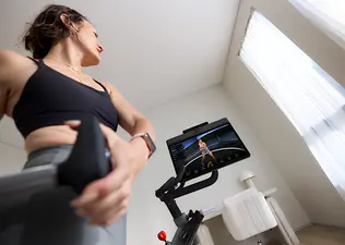 Woman lifts weights during a cross training workout for cycling while standing next to a Peloton Bike+.