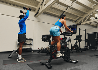 Two friends doing strength and cardio workouts side by side in a gym using a Peloton Bike.
