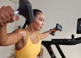 Woman doing a strength workout using dumbbells and a Peloton Cross Training Series treadmill wondering when she will see results from working out.