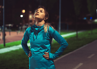 An exasperated athlete on an outdoor running path in the evening, with her hands on her hips and head pointing up to the sky. She has lost motivation to work out.