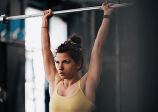 Young woman in a yellow tank top performing an overhead press with a barbell in a gym setting