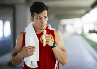 An athlete eating a banana after a workout while walking outside. He is wearing a red athletic tank top and has a white sweat towel over his shoulder.