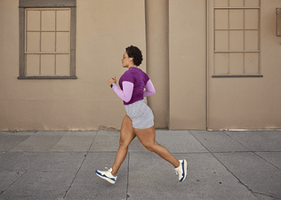 Woman in a purple shirt and grey shorts doing the run-walk method outside past a blank wall