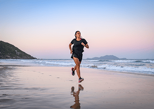Woman running on the beach at sunset in sneakers and a black outfit.