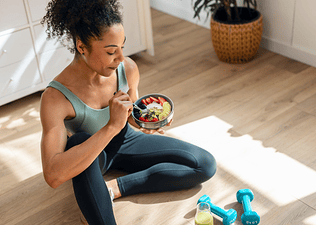 An athlete sitting on the floor at home eating a fruit and yogurt bowl after finishing a dumbbell workout.