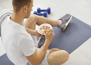 A man eating oatmeal and fruit with an optimal carb-to-protein ratio on a yoga mat after finishing a workout.