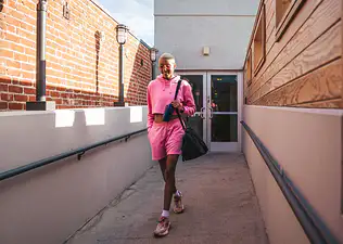 A young woman leaving the gym. She's wearing a pink outfit set with a gym bag on her shoulder. 