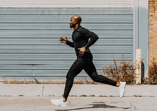 A man on a long run outside. He's wearing all black and jogging in front of an industrial building.