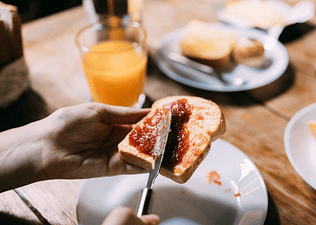 A person spreading jam on a piece of toast next to a glass of orange juice. This meal would make an ideal breakfast before a marathon.