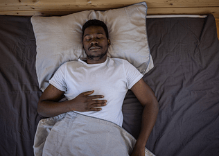 A man practicing breathing exercises for sleep. He's lying on his back in bed, eyes closed,  with his right hand on his upper abdomen.