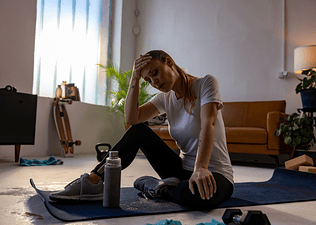 A woman sitting down on a yoga mat at home with her head resting in her hands and her eyes clothes as she takes deep breaths. She felt dizzy when stretching.