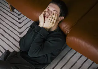 A man leaning against a couch at home while rubbing his eyes as he deals with the Sunday scaries.