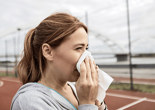 In this image for an article about whether or not you should exercise when you're sick, a woman is blowing her nose into a tissue while she stands outside on a sports court of some kind.