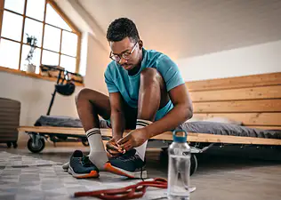A man practicing self-discipline for exercise. He is sitting on his bed and tying his workout shoes before exercising, even if he may not feel like it. 