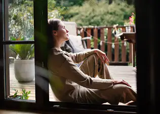 A woman sitting down at home and leaning against a doorway as she looks off into the distance outside.