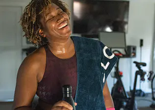 A woman laughing and smiling after a workout. She is holding a water bottle and has a towel draped around her left shoulder.