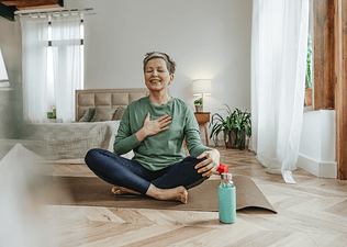 A woman meditating on an indoor yoga mat, with one hand on her chest and the other on her knee, to practice self-care during menopause.