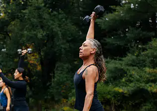 A middle-aged woman lifting a dumbbell in the air while doing an outdoor workout.