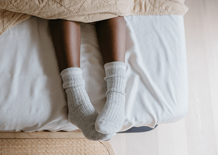 A close-up photo of a woman sleeping with socks. Her socks are gray and her feet are popping out from under the covers on her bed.