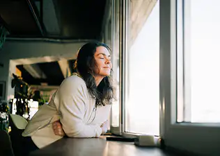 A woman leaning on a counter near a window, closing her eyes and taking time to be mindful.