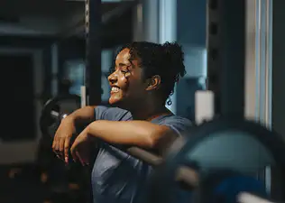 A woman smiling and resting her arms on a barbell. She is enjoying her exercise routine and views working out as a hobby.