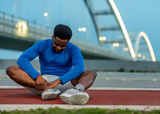 A male athlete sitting on the ground and holding his right ankle. He's experiencing ankle pain after running.
