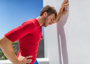 A tired male athlete leaning against a white wall outside. His face is red, with one hand on his hip and another arm leaning against the wall. He's resting his head against his arm and wearing a red T-shirt. It's a blue, sunny day outside.