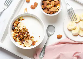 An array of high-protein snacks on a white table, such as Greek yogurt, bananas, and nuts.