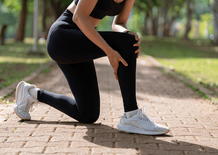 Close-up photo of a runner bent down on one knee on an outdoor path, scratching their calf and holding their knee.