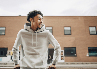 A man in a hoodie smiling and leaning against a railing outside. He's looking to his left.