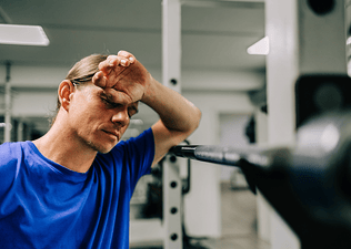 A tired man in the gym near a barbell. His hand is covering his face and his eyes are closed. He's wearing a bright blue shirt.