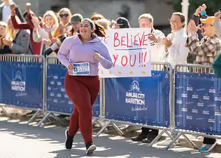 A woman happily running a marathon while a spectator behind her holds up a marathon sign that says "We believe in you!"