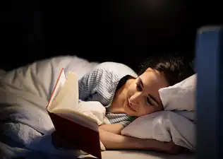A woman reading a book while lying in bed at night as part of her bedtime routine.
