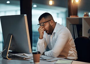 A man who only got six hours of sleep is yawning and rubbing his face while sitting at his work desk.