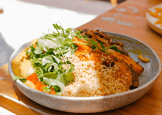 A high-protein grain bowl featuring quinoa, leafy greens, and pumpkin. The bowl is sitting on a wooden table in the sunlight.