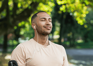 A man practicing mindfulness and meditation. He is closing his eyes and smiling while listening to wired headphones in an outdoor setting.